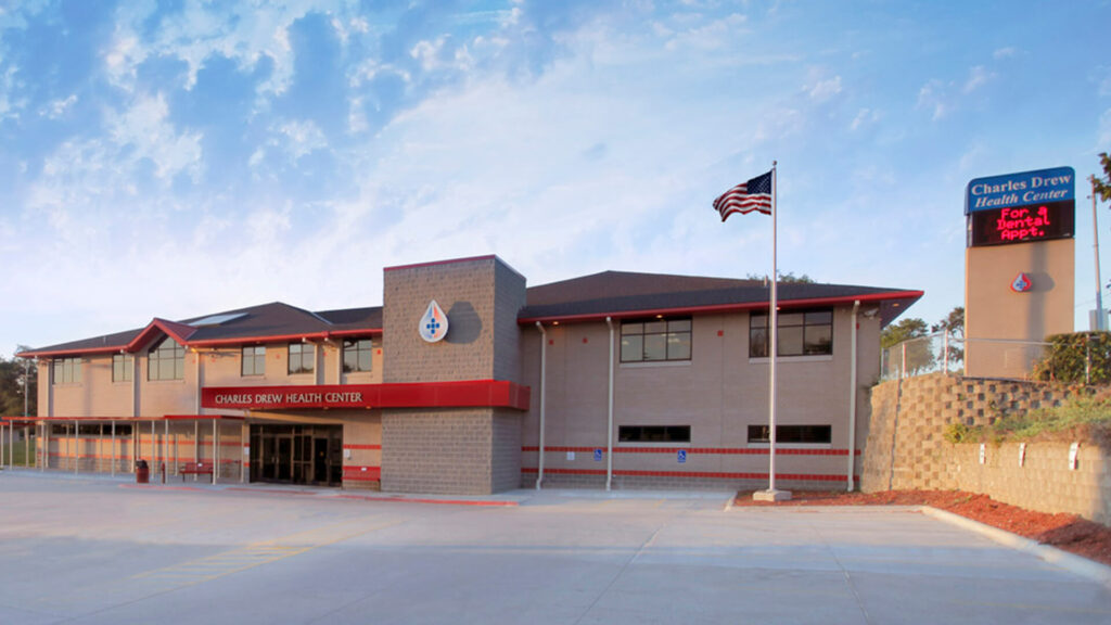 Exterior of a beige building with signage that reads Charles Drew Health Center. The parking lot is empty and we see an American flag waving on a flag pole beside the building. The sky is blue and cloudy.