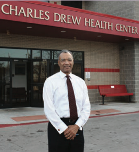 An African American man stands in a white shirt and tie in front of Charles Drew Health Center.