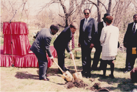 A group of diverse individuals watch as two men holding shovels begin digging into the ground at a groundbreaking event.