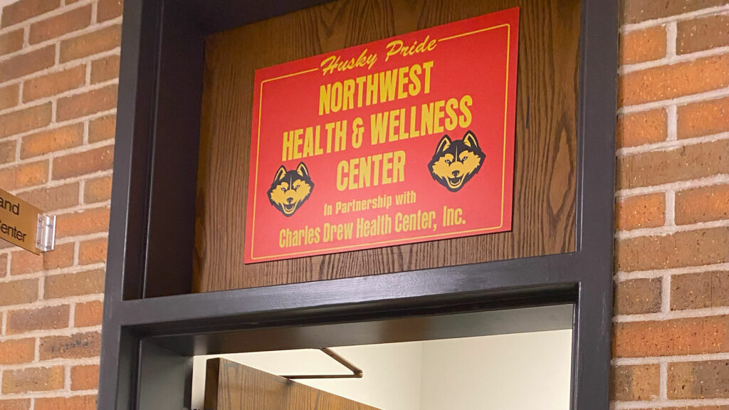 A red sign over the doorway inside of a school building reads Northwest Health & Wellness Center in partnership with Charles Drew Health Center. Two husky dog mascot illustrations flank the text on the sign.