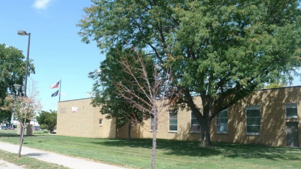 A big, green tree stands outside the exterior of Kellom Elementary School. The building is made of tan brick and the sky is bright blue.