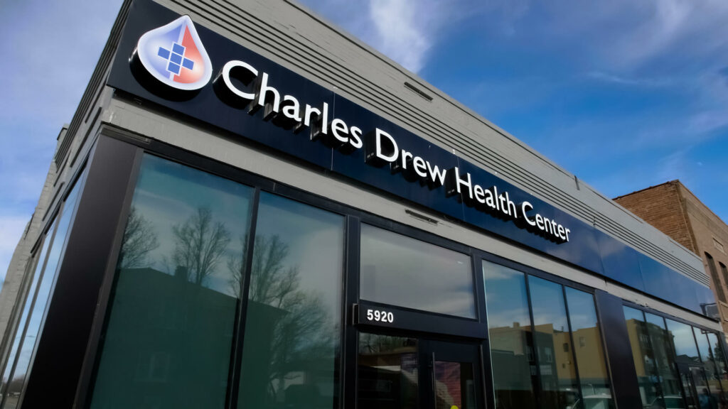Close up on signage on the exterior of a grey brick building with many windows. The signage reads Charles Drew Health Center.